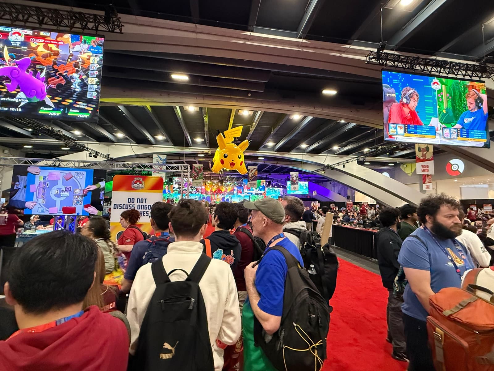 A busy convention floor with screens, a Pikachu figure and crowds moving between stands