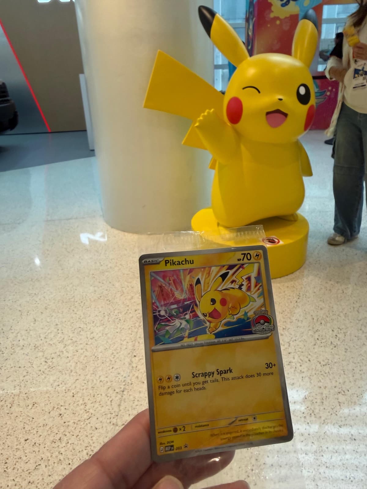 A hand holding a Pikachu promo card in front of a large Pikachu statue